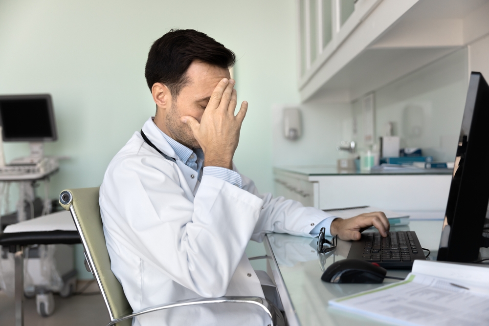 Doctor sit at desk with computer and papers, cover face with hand, feel exhausted